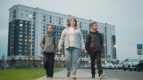 woman confidently walks hand in hand with two children in urban neighborhood near tall apartment buildings during evening, symbolizing parental care, unity, and support in daily life with school backpacks