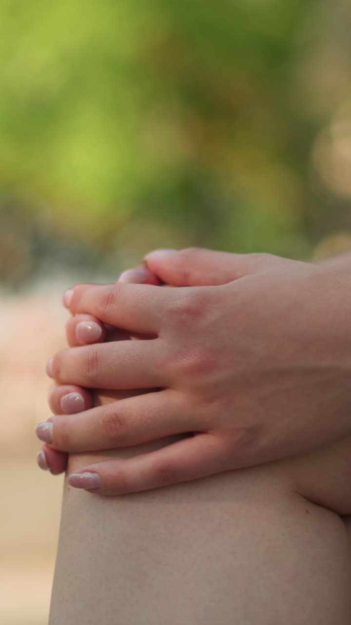 primer plano de una mujer joven sentada al aire libre con las piernas cruzadas, las manos juntas descansando en la rodilla, usando un brazalete de mano negro, bajo luz natural suave con fondo verde borroso