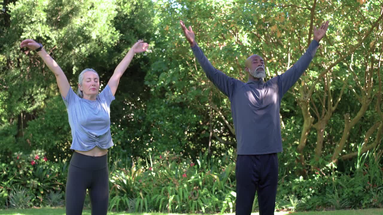 Coppia multietnica che pratica yoga in piedi su un prato con un dispositivo da polso, iniziando con le mani unite sopra la testa per respirare.