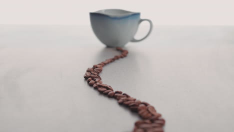 Lined-Roasted-Coffee-Beans-Towards-Coffee-Cup-Against-White-Surface