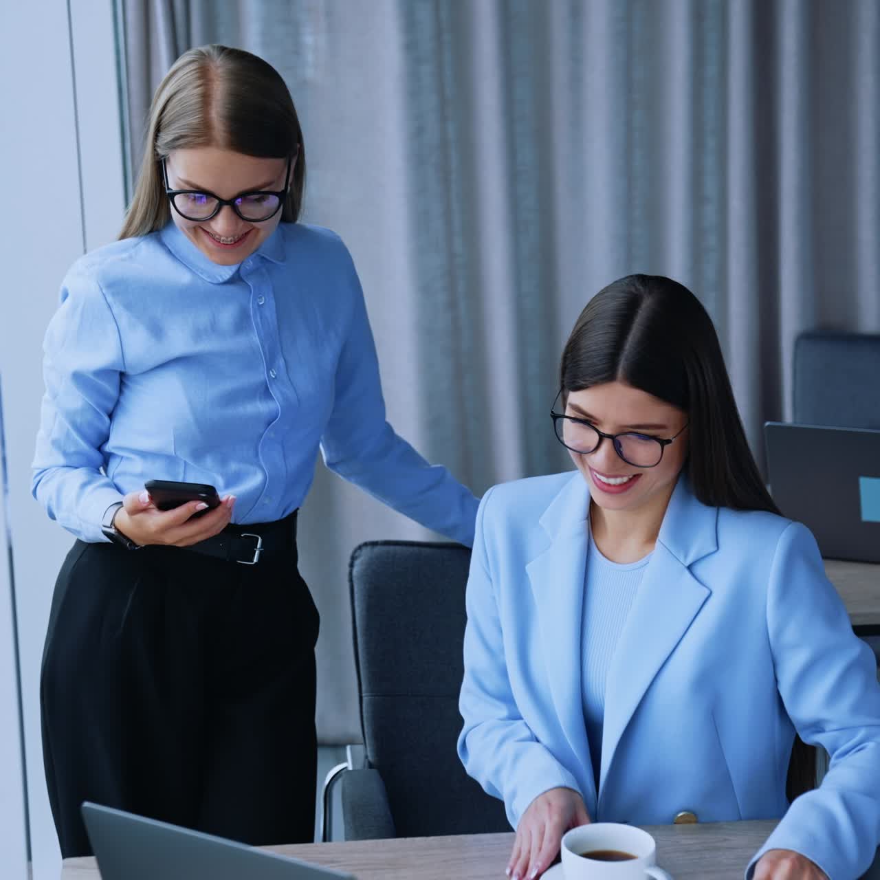 Female colleagues communication at work. Long-haired ladies in glasses talk, laugh and look at laptop. Brunette woman having cup of coffee