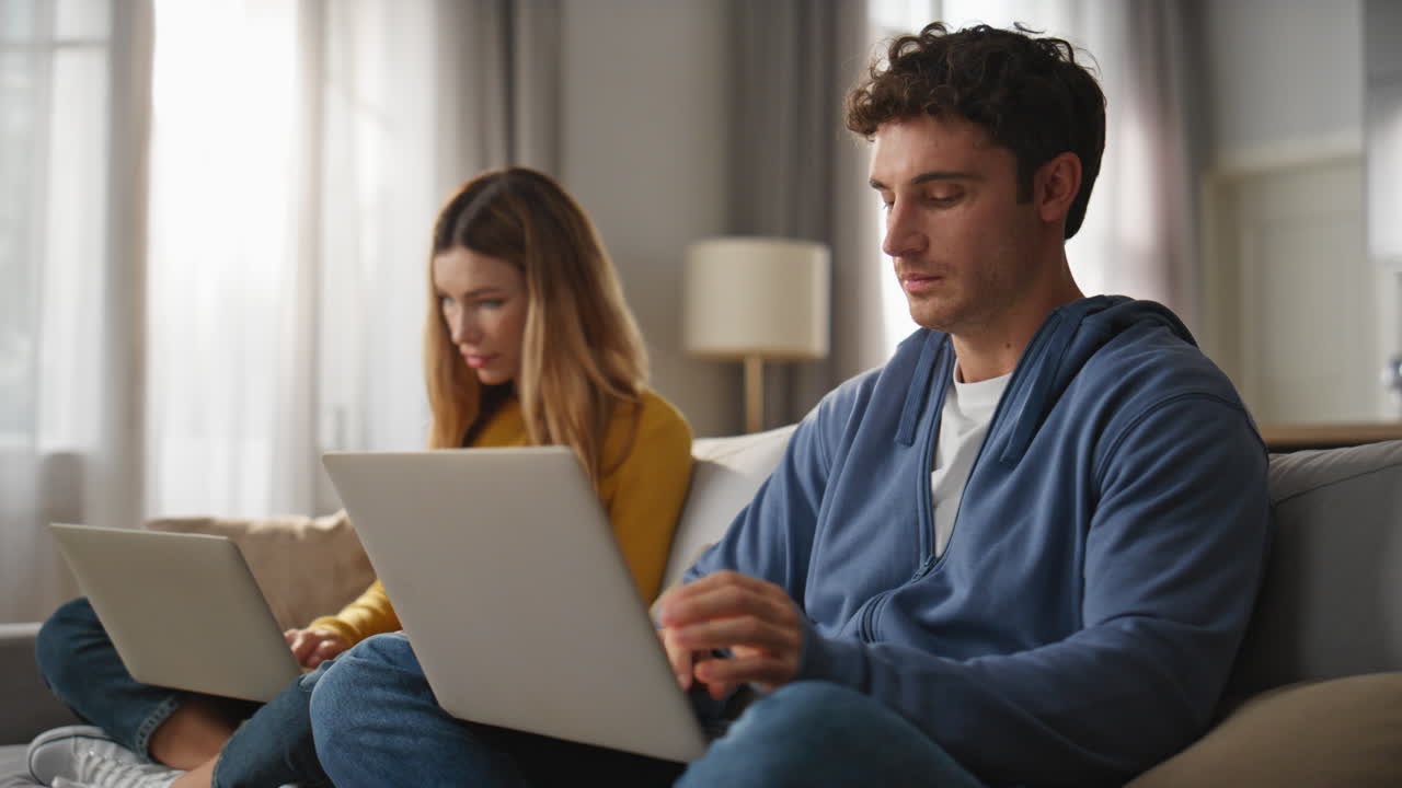 Focused spouses working computers couch together closeup. Young pair freelancers