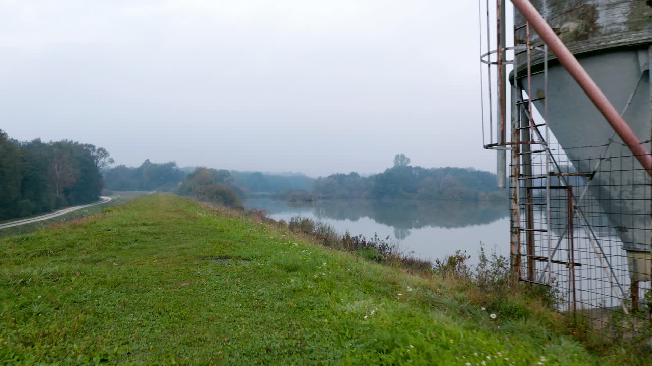 toma panorámica de un silo de metal en una colina cubierta de hierba con vistas a un lago transparente y reflectante en una brumosa mañana de otoño