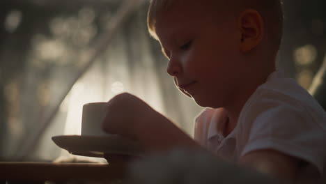 child drinks hot beverage from cup at home. dreaming boy turns cup of tea sitting at table in morning closeup. cute preschooler has breakfast