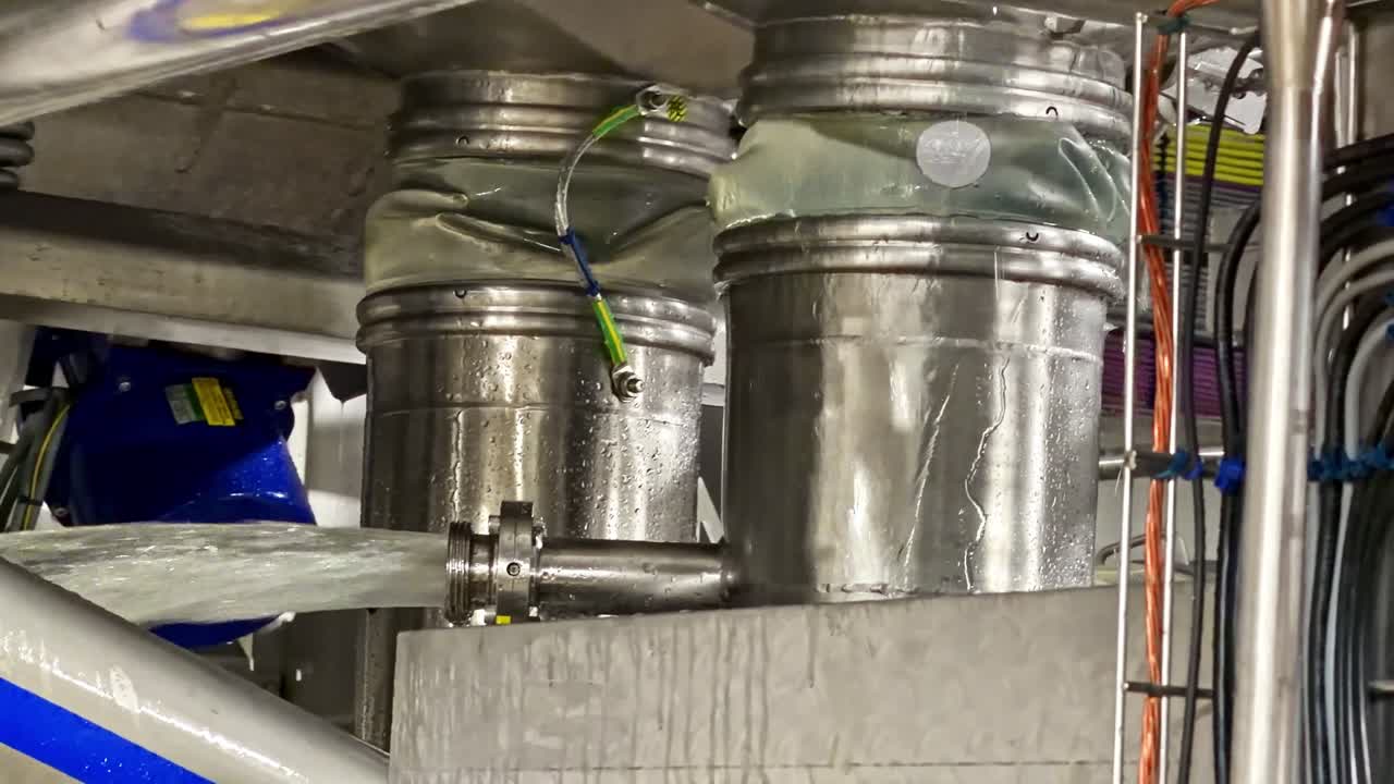 Close-up view of water pouring from stainless steel hoppers with plastic bags and hoses into connected piping system in food or dairy processing facility