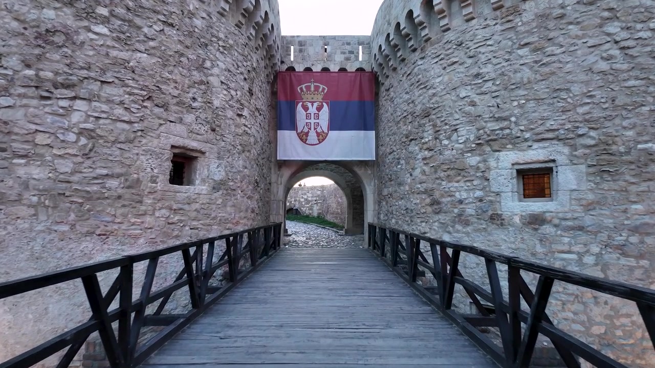 I visitatori camminano lungo il ponte di legno verso l'ingresso di un castello. Le mura sono in pietra. La bandiera serba è appesa sopra l'arco. L'ambientazione è serale.