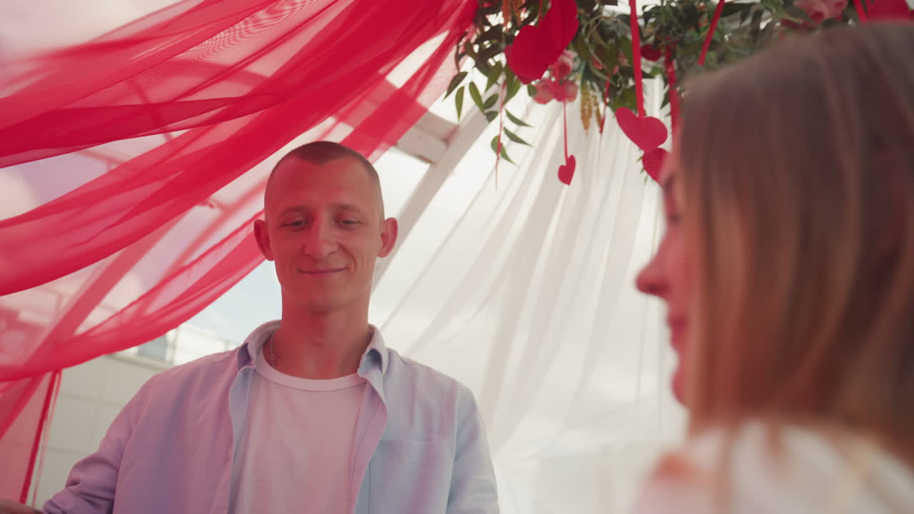 coppia caucasica sotto un baldacchino rosa che ammira fiori sospesi e si tiene per mano mentre l'uomo offre una rosa, drappeggi rossi trasparenti filtrano la calda luce del sole, sorrisi spontanei, donna bionda con tatuaggio, festeggiamenti rilassati