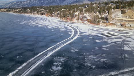 Overhead-aerial-drone-view-of-Windermere-Lake-frozen-over-passing-along-the-southeastern-shoreline-with-ice-skaters-and-manmade-rinks-made-on-the-ice-below