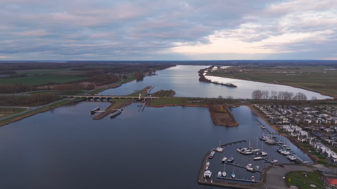Aerial view of a river, bridge, and marina at sunset, with golden light reflecting on the water and a forested shoreline. Dutch landscape.