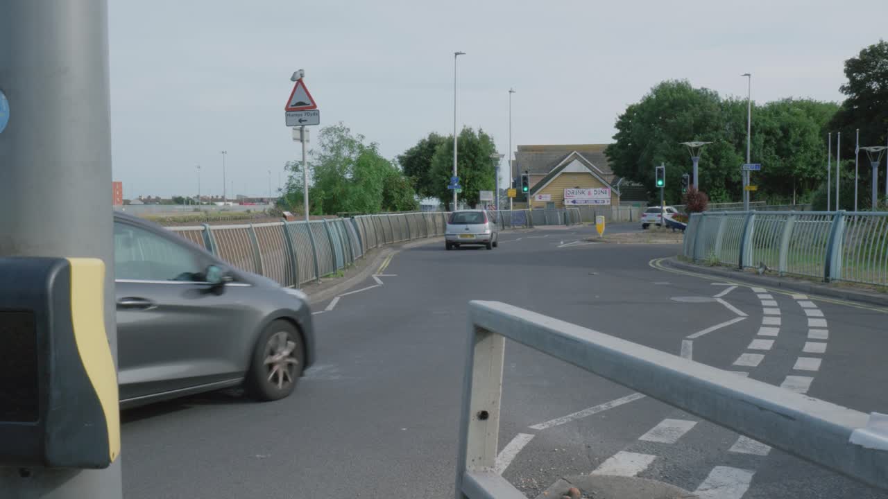 A pedestrian crossing with a silver car waiting. Traffic signs, street lamps, trees, and buildings are visible in the background. Roundabout with white lines and railings.Yellow button for pedestrians