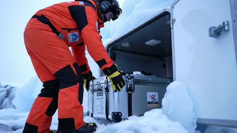 an arctic researcher in an orange suit carefully loads a high-tech case into a storage unit surrounded by ice, showcasing the challenges of scientific expeditions in extreme environments
