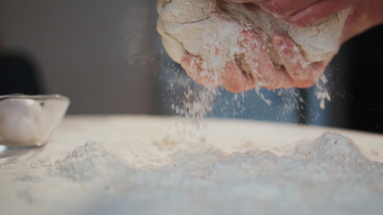 Close-up de mãos colocando delicadamente a massa em uma superfície enfarinhada. Mãos levemente polvilhadas com farinha, mostrando um toque cuidadoso. A massa parece macia e maleável, repousando sobre pó branco. Ideal para tutoriais de culinária