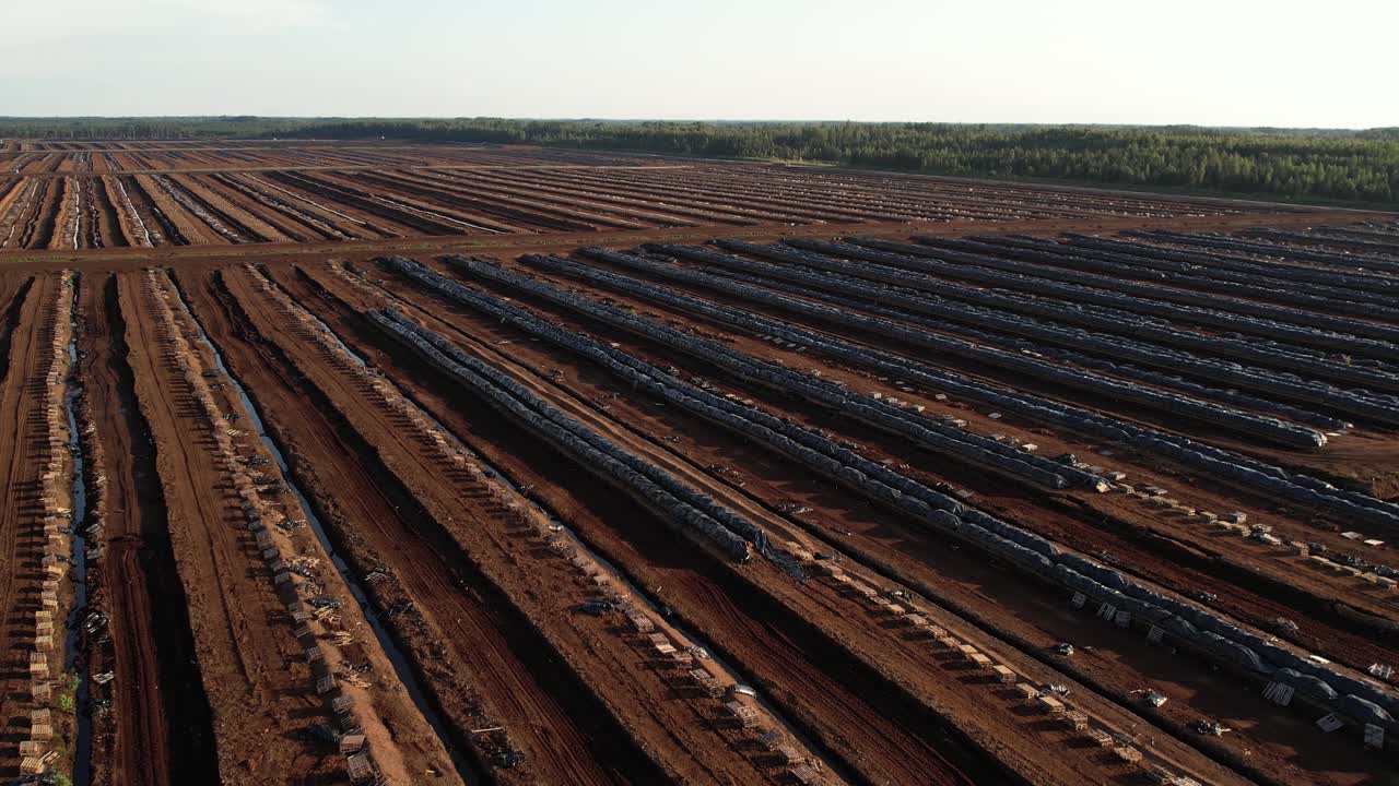 Aerial view of a large peat bog harvesting site with long parallel rows of extracted peat, wooden pallets, and covered stacks stretching across the landscape