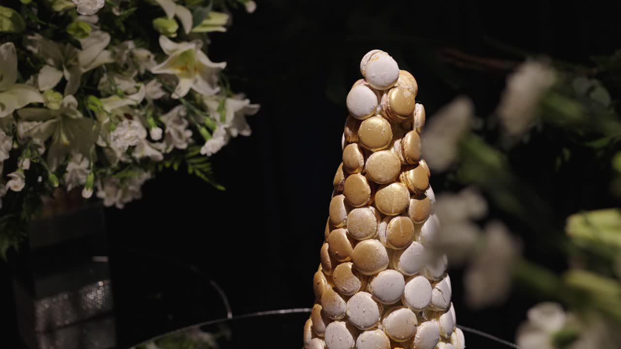 Pyramid of assorted macarons arranged as decorative centerpiece on luxury wedding dessert table with floral background and soft ambient lighting, shallow depth of field, slow motion reveal shot