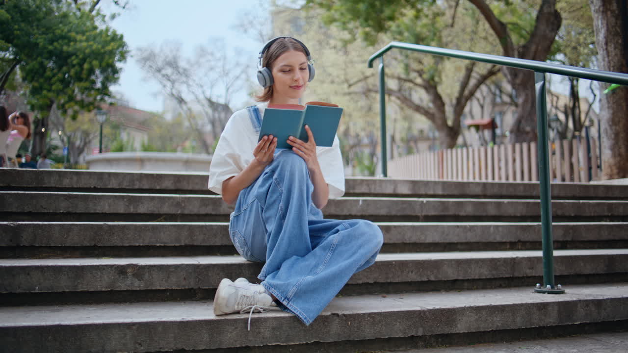 Giovane hipster felice si gode un libro seduto sui gradini in strada con le cuffie. Donna spensierata