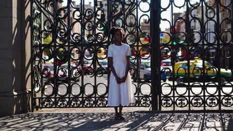young woman in a white dress stands serenely at an ornate wrought iron gate, the bustling city street visible through its intricate design, symbolizing the intersection of sacred and secular life