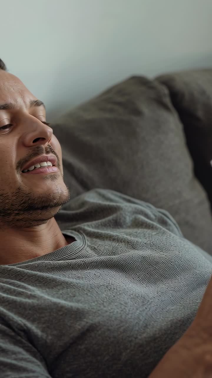 Video verticale: Uomo sorridente con maglietta grigia che reagisce al telefono che si sposta e si adagia sul divano grigio.