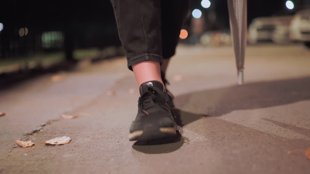 Close up of legs wearing black shoes and cropped pants walking slowly with umbrella on quiet outdoor path scattered with fallen leaves under soft street light during calm peaceful night atmosphere