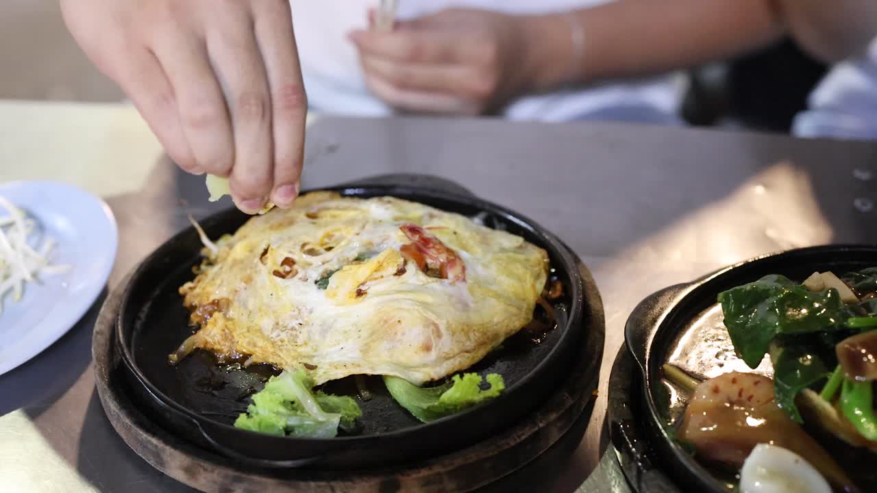 Hand sprinkles seasoning onto hot Thai omelette, vibrant street food setting, natural lighting, overhead view