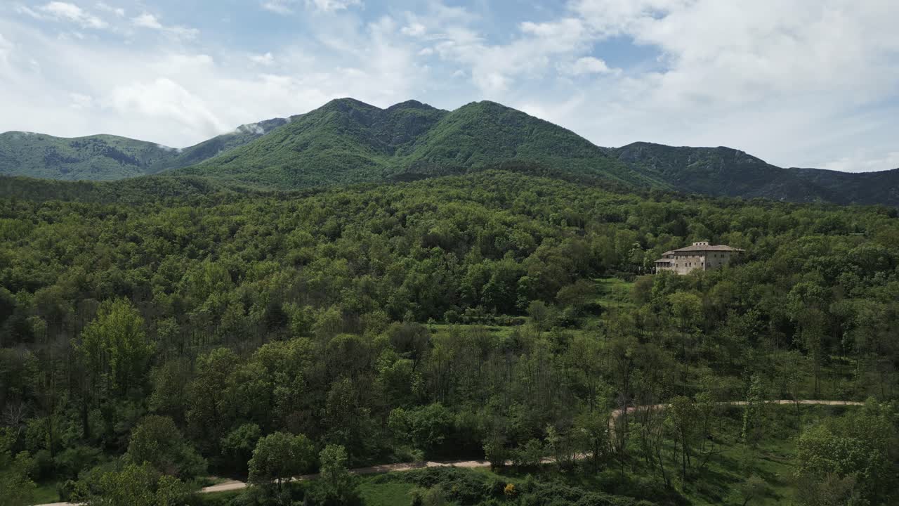 Green mountain landscape revealing lush forest surrounding isolated building, winding road leading through verdant terrain under overcast cloudscape