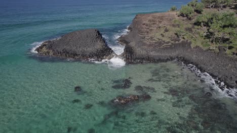 waves splashing on hexagonal volcanic rock formations with surfer in the sea revealed