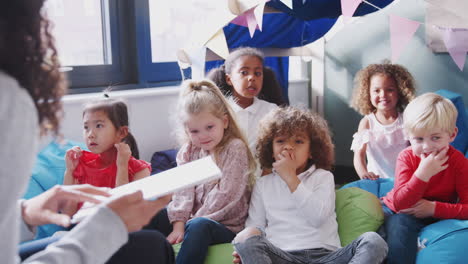 female infant school teacher showing a book to kids in a comfortable corner of classroom, close up