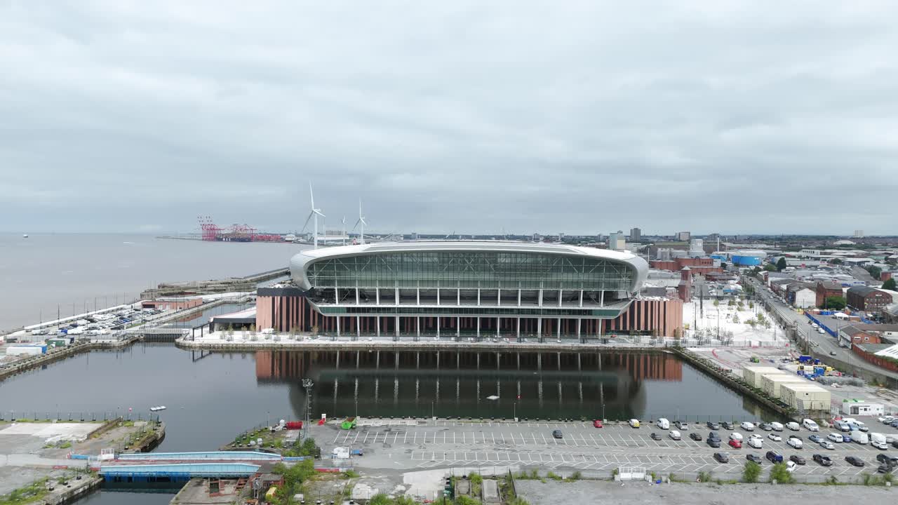 Modern architecture of the Hill Dickinson Stadium, reflecting on the calm waters of the dock, with wind turbines and cranes visible in the distance