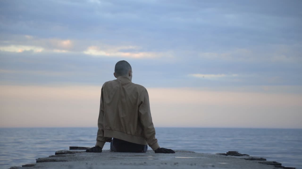 Back View Of A Man Sitting Alone On Wooden Jetty At The Beach Under A Cloudy Morning Sky - Medium Shot