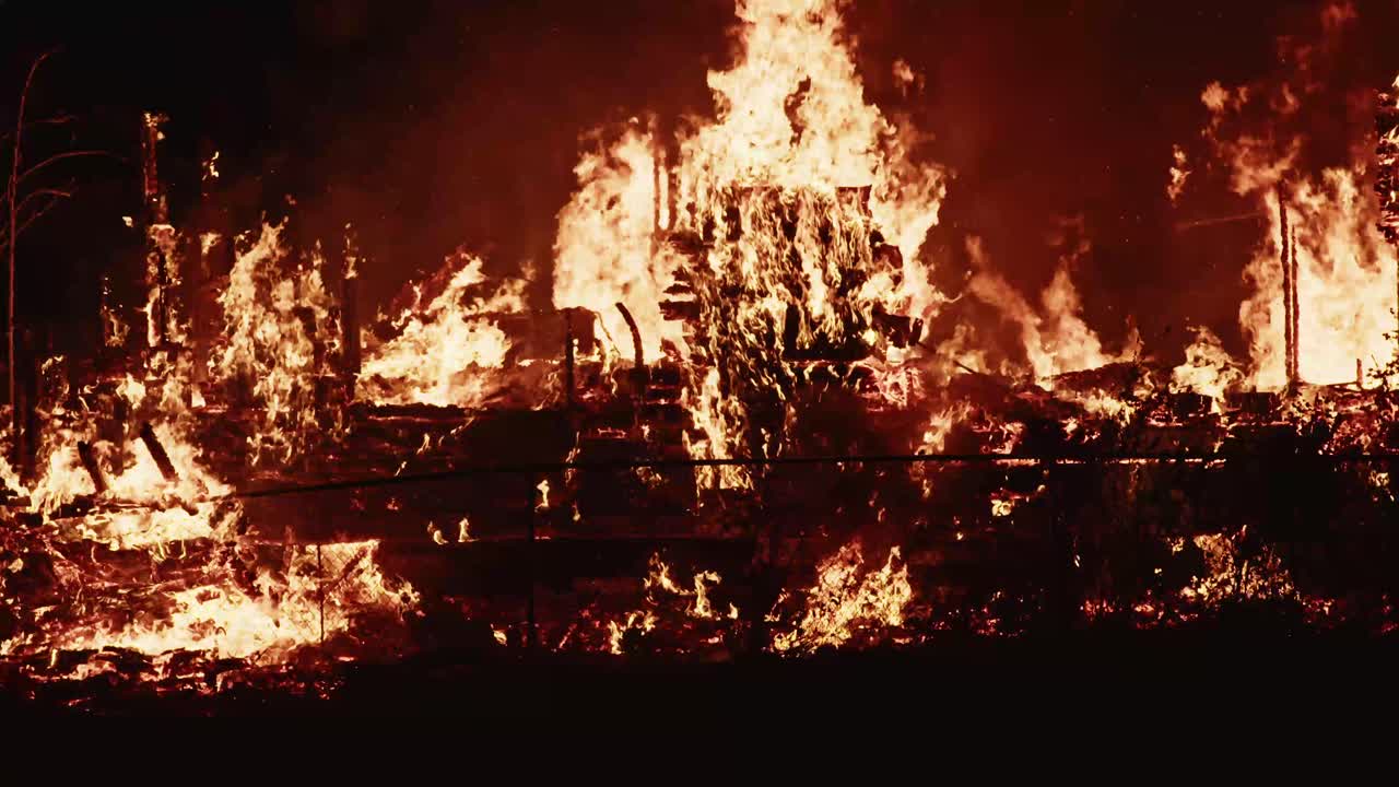 Massive nighttime blaze consumes a structure, with flames and smoke erupting behind a chain-link fence in a dramatic fire scene