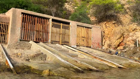 Old-fishing-boat-sheds-by-the-sea-in-Ibiza-Spain,-traditional-fishing-huts-at-a-sunny-beach,-boat-garages-on-the-shore-with-turquoise-water,-4K-panning-right