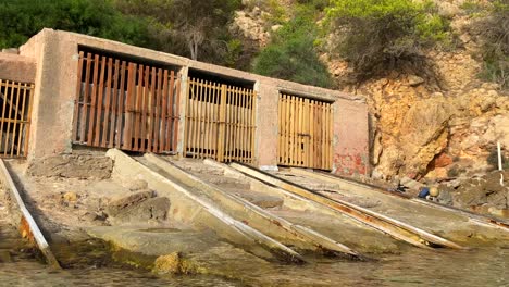 viejos cobertizos de barcos de pesca junto al mar en ibiza españa, cabañas de pesca tradicionales en una playa soleada, garajes de barcos en la orilla con agua turquesa, 4k panorámica a la derecha