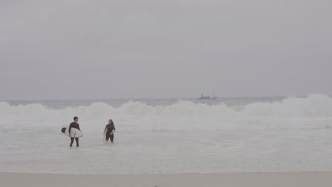 Two surfers walking through waves on a cloudy day