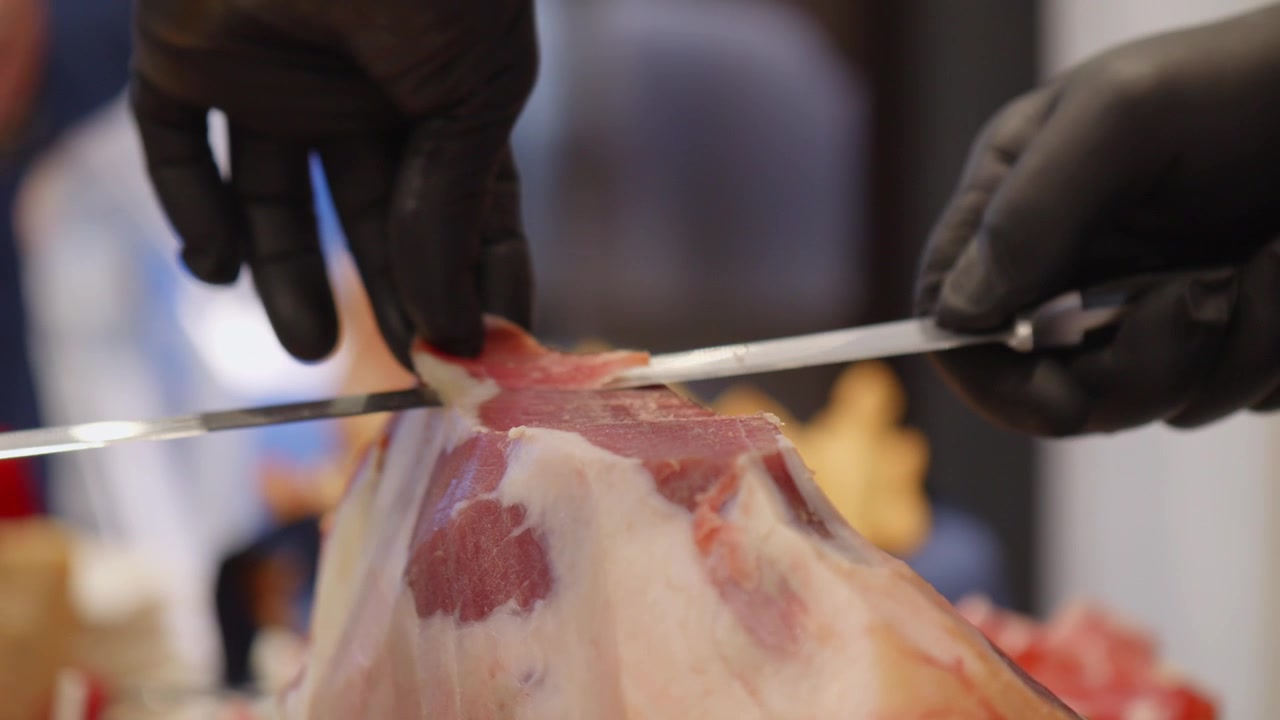 Närbild på en kock som försiktigt skär tunna skivor från en hel prosciutto crudo med en lång trancherkniv och svarta handskar.