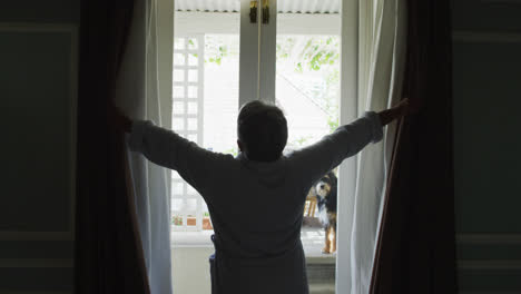 vista trasera de una mujer de raza mixta senior en casa abriendo cortinas