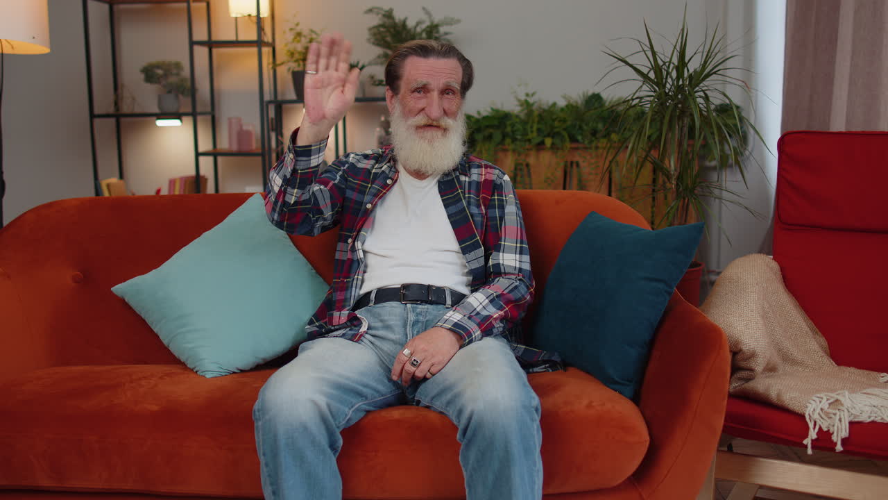hombre mayor sonriendo amigablemente a la cámara, agitando las manos gestando hola, hola, saludo en casa en el sofá