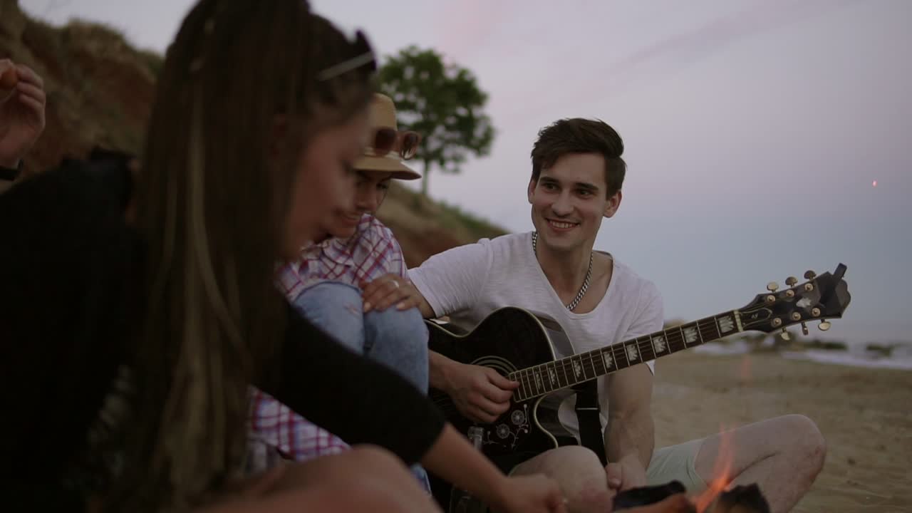 Young friends sitting by the fire on the beach in the evening, grilling sausages, drinking beer and playing guitar. Slow Motion