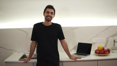 portrait of young confident caucasian man standing in kitchen, smiling, looking at camera, full of optimism, enjoying his life, dark brown hair, brown eyes. laptopn on a counter