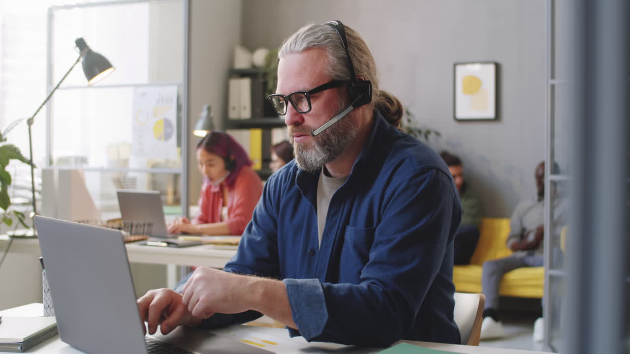 Senior Man in Headset Using Laptop and Talking on Web Call in Call Center