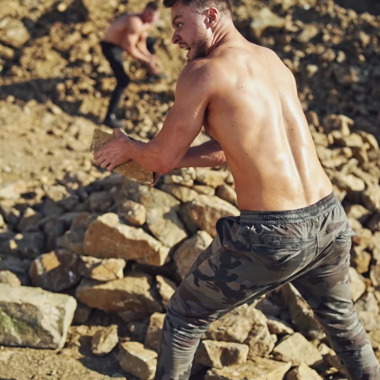 Men exercises by throwing stones in a sunny day. Two shirtless sportsmen doing workout on the rocks background. Slow motion.