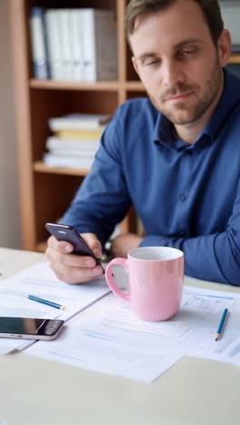 a man is sitting at a desk with a pink mug and a cell phone
