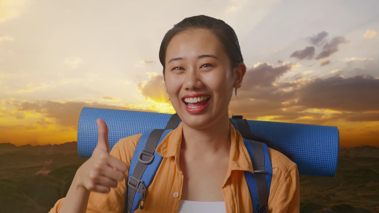 Close Up Of Asian Female Hiker With Mountaineering Backpack Smiling And Showing Thumbs Up Gesture To Camera While Standing On The Top Of Mountain During Sunset Time