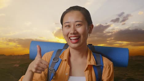 close up of asian female hiker with mountaineering backpack smiling and showing thumbs up gesture to camera while standing on the top of mountain during sunset time