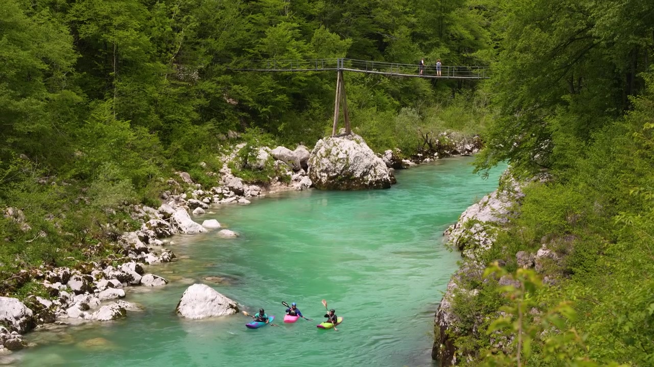 Three kayakers paddle down turquoise river through lush forest with footbridge above