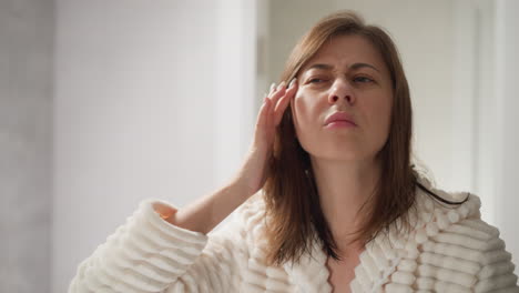 mujer en túnica mira la cara en el espejo después de ir al centro de spa. atractiva dama con cabello oscuro mira el efecto después de la máscara facial en el área de spa espaciosa