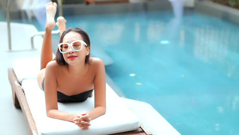 a pretty woman enjoys her time poolside by lying prone on a deck lounger while playfully kicking her feet