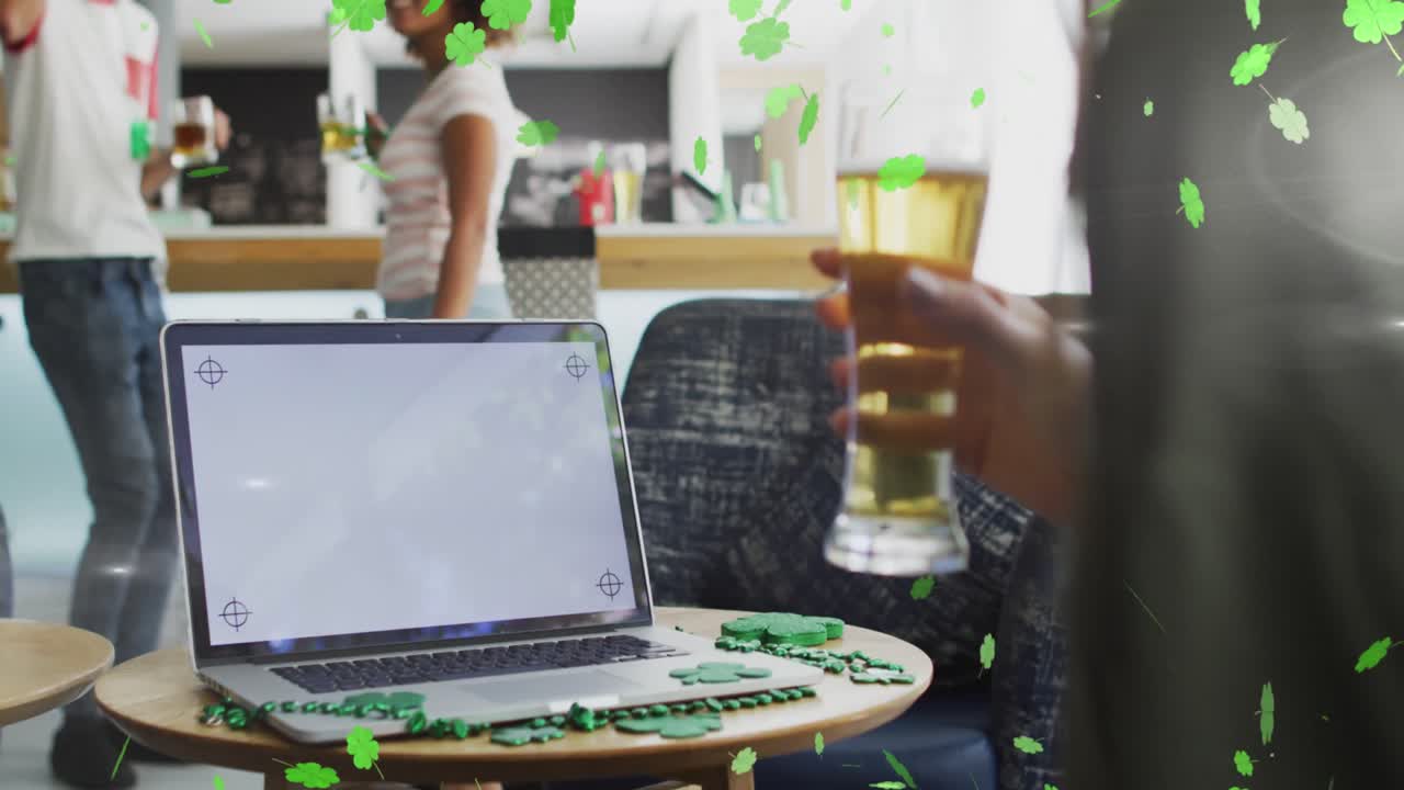 Animation of clover icons over diverse friends drinking beer and using laptop with copy space