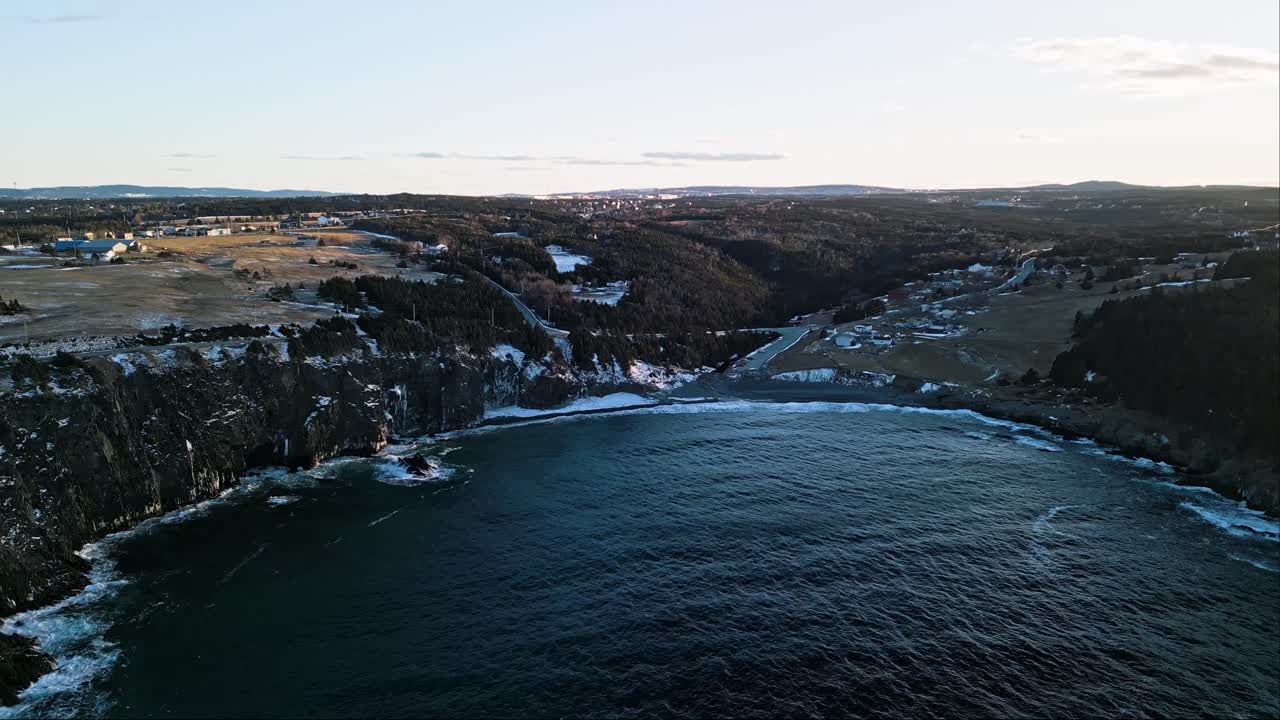 A drone swoops over Middle Cove's rugged coastline in Newfoundland, revealing dramatic sea cliffs and Atlantic waves crashing against the rocks under a pastel sunset sky