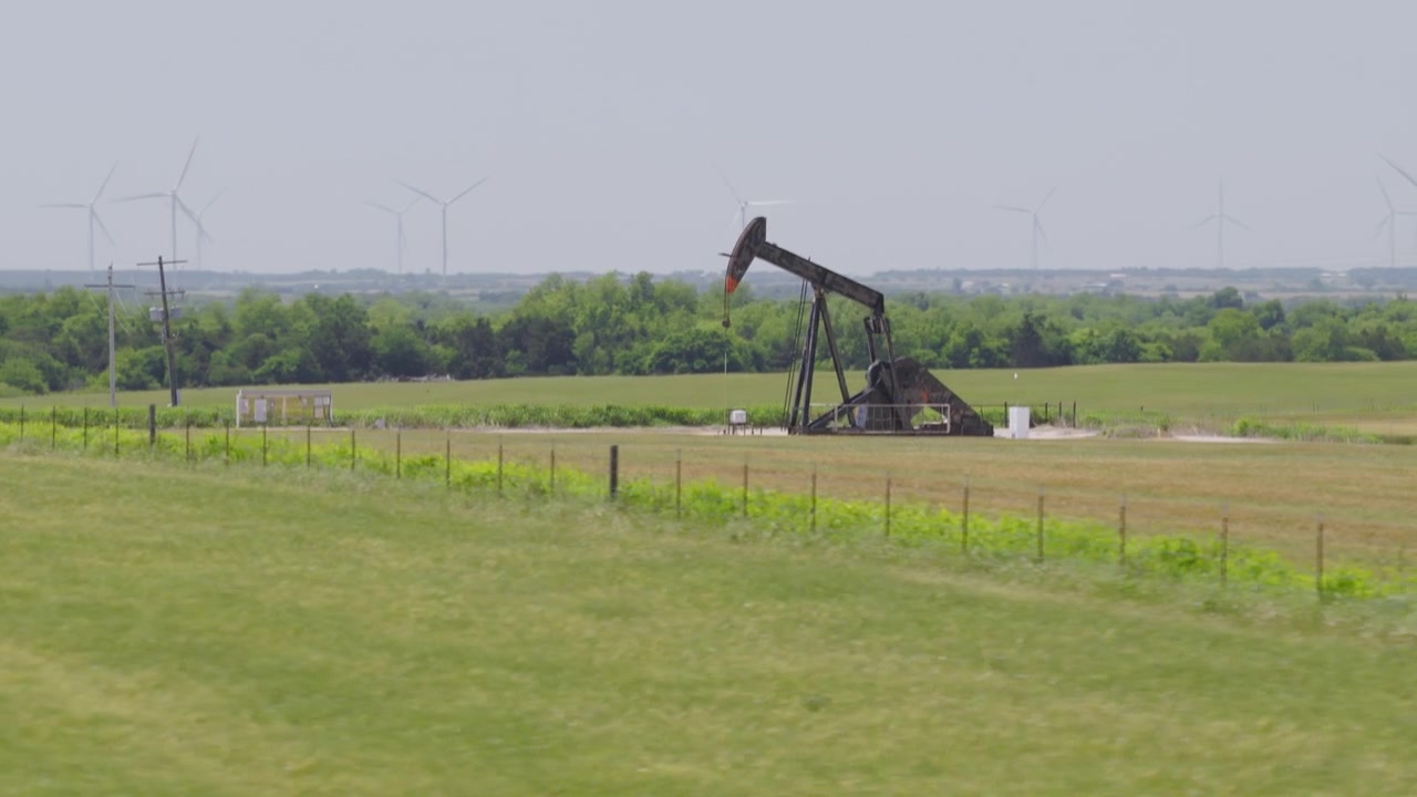 Drone 4K of a green field with an oil pump jack and wind turbines in the background.