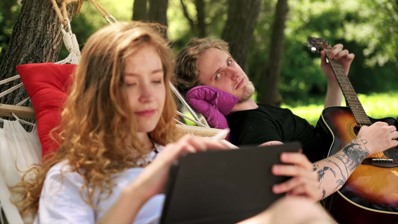 Couple lying on hammocks. Young man is playing guitar while woman is using tablet with stylus. Greenery around. Glamping. Slow motion