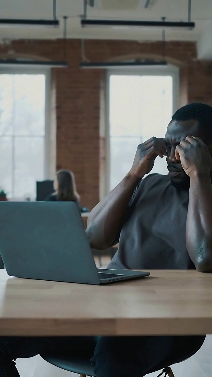 Vertical video: Rubbing temples, man in dark tee staring at open laptop at open office, copy space
